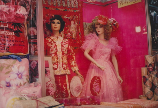 A color photograph of two White Barbie-like dolls, one wearing an Asian-style red and gold outfit and the other a pink gown, in a shop window.