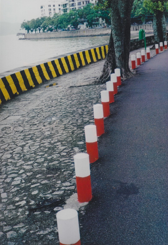 A color photograph of red and white bollards at the edge of a street, a stone sidewalk, and a yellow and black temporary floodwall.