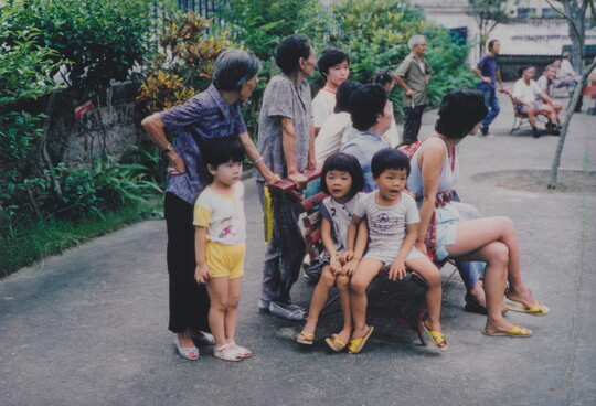 A color photograph of Asian children and adults sitting on and standing around a bench on a sidewalk.