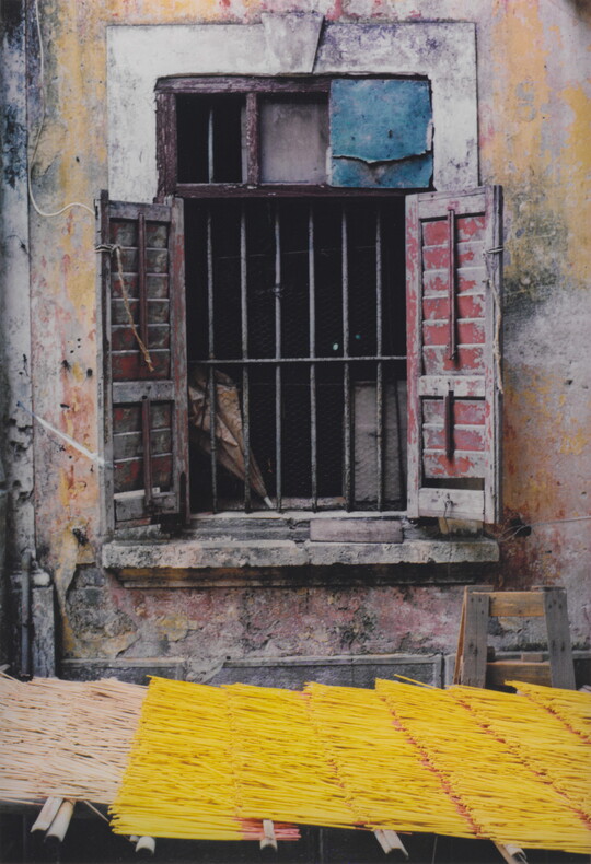 A color photograph of a barred window, wood shutters open, set in peeling yellow plaster above a yellow thatched awning.