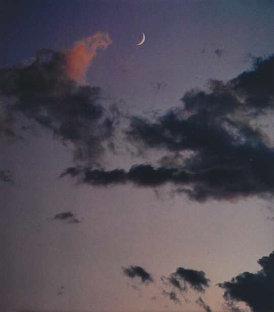 A color photograph of a crescent moon at dusk above thin, dark clouds.