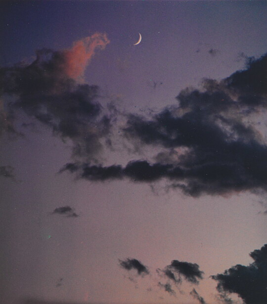 A color photograph of a crescent moon at dusk above thin, dark clouds.
