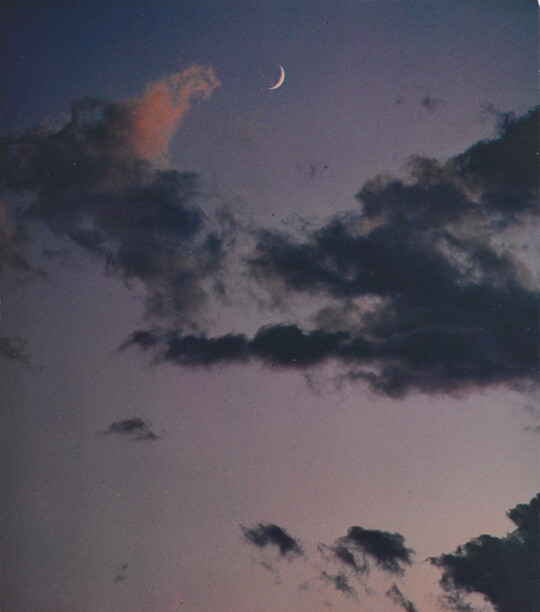 A color photograph of a crescent moon at dusk above thin, dark clouds.