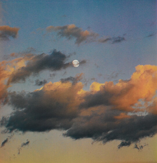 A color photograph of a full moon at dusk behind wispy, orange and dark clouds.