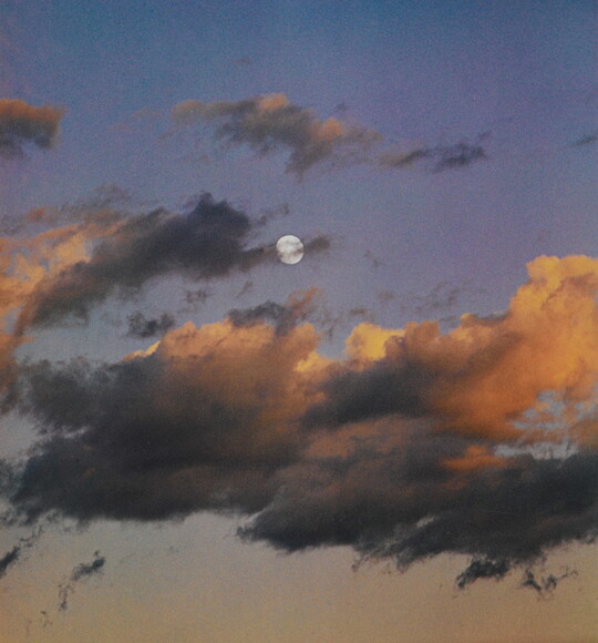 A color photograph of a full moon at dusk behind wispy, orange and dark clouds.