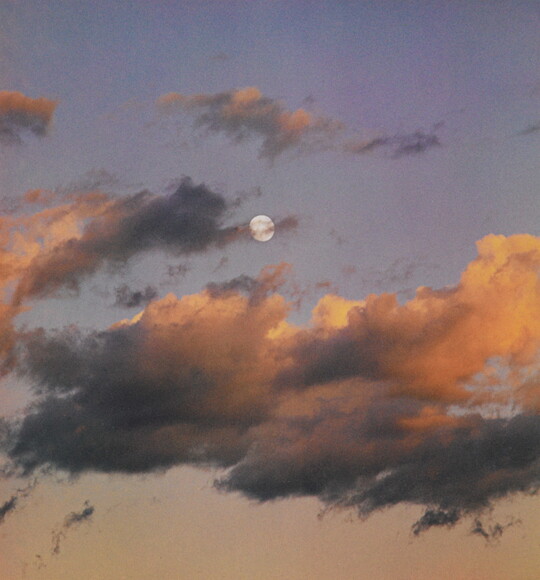 A color photograph of a full moon at dusk behind wispy, orange and dark clouds.