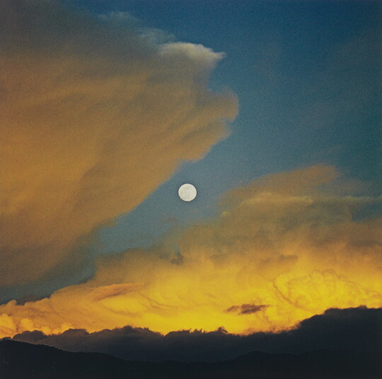 A color photograph of a full moon in a dark blue sky with yellow swirling clouds.