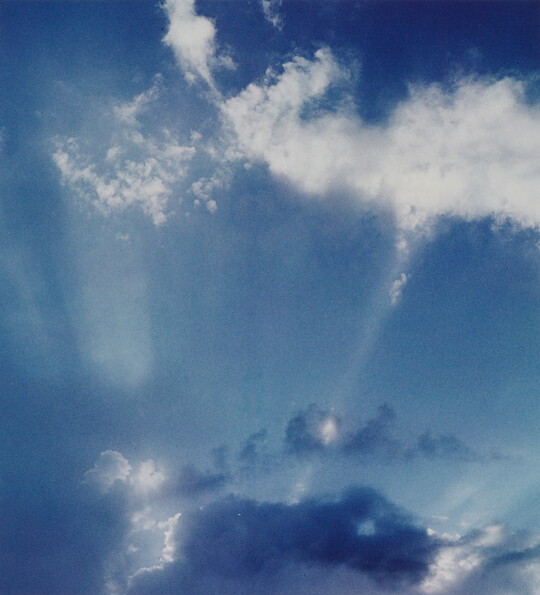 A color photograph of white clouds in a blue sky and sun rays that create white streaks.