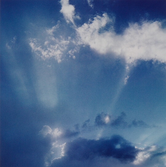 A color photograph of white clouds in a blue sky and sun rays that create white streaks.