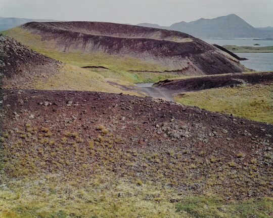 A color photograph of a crater on a rocky and green landscape with water and mountains in the distance.