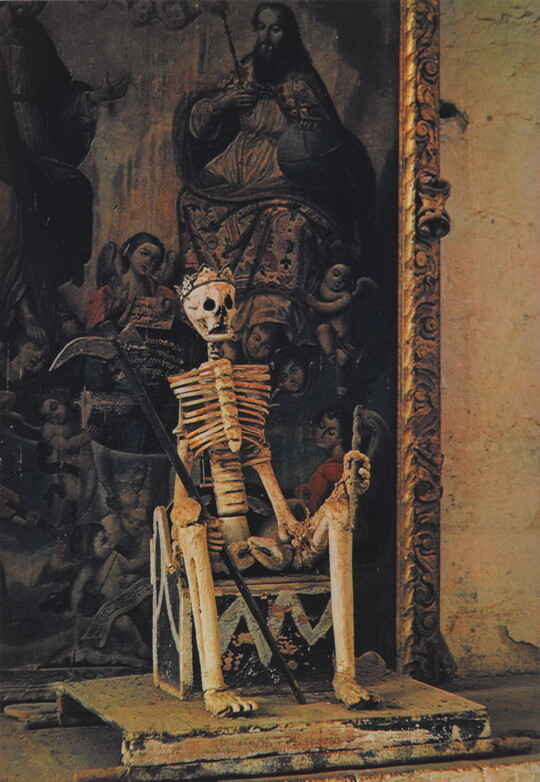 A color photograph of a skeleton seated on a throne, wearing a crown and holding a scythe in one hand, in front of a painting.