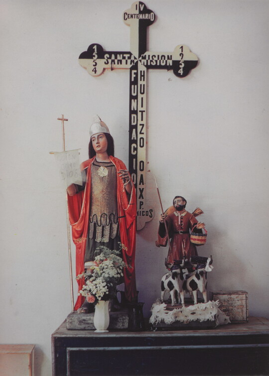 A color photograph of a table with figures of a saint in a red robe holding a cross staff, and a cowherd with two cows; a large black-and-white cross in the background.
