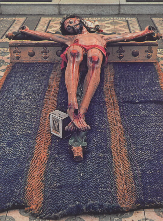 A color photograph of a painted sculpture of a crucified Jesus, eyes closed, wearing a crown of thorns; the entire crucifix laying on a blue cloth on a tile floor.