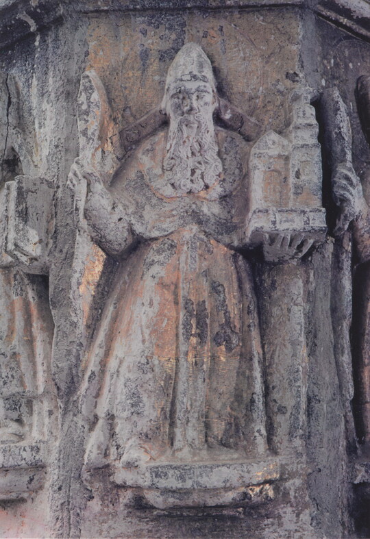 A color photograph of a stone figure with a long beard, wearing robes, holding a building in one hand and what might be a staff in the other.