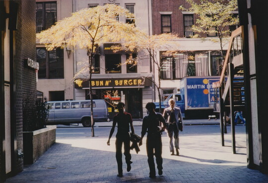 A color photograph of three men walking in an alley paved with bricks; behind them are small trees lining the sidewalk and a restaurant on the opposite side of the street.