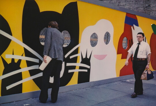 A color photograph of two men walking past a construction fence decorated with a cartoonish black cat, ghost, and other characters; one man peeks through the fence.