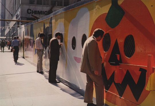 A color photograph of men in business attire on a city sidewalk looking through holes in a construction wall that is painted with a mural of pumpkins and ghosts.