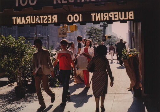 A color photograph of a busy sidewalk, several people walking under an awning advertising a restaurant.