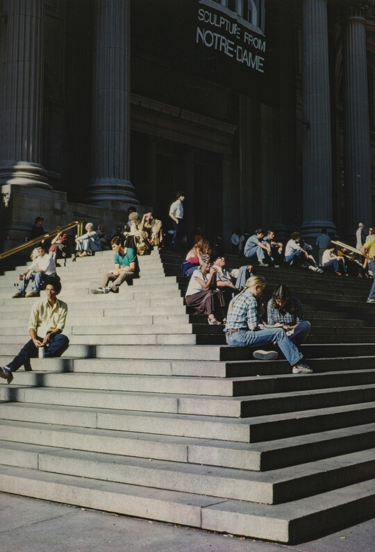A color photograph of people sitting on the stone steps of a building with large fluted columns.