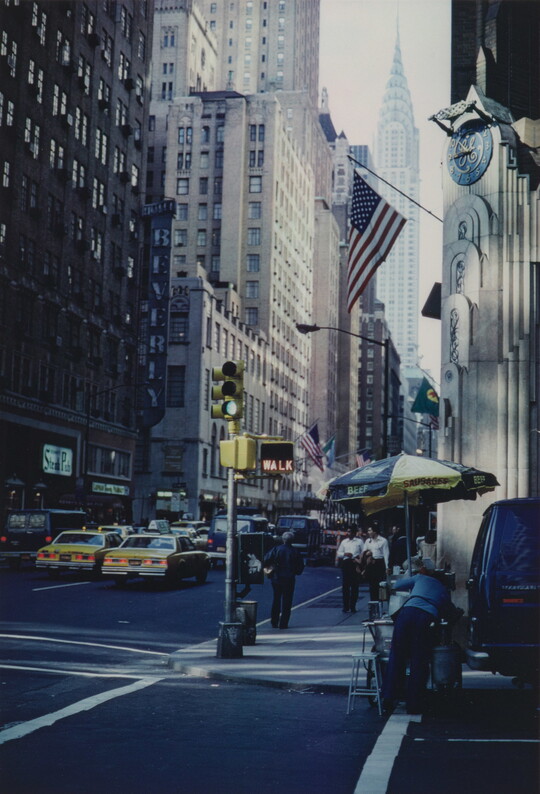 A color photograph of a busy city street with taxis, pedestrians, a hot dog cart, and the Empire State Building in the background.