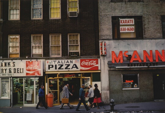 A color photograph of pedestrians walking past storefronts advertising "Italian Pizza" and "Coca-Cola."