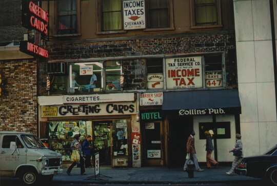 A color photograph of the front of a building advertising a greeting card shop, a hotel, a pub, and other services as people walk past.