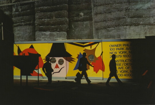 A color photograph of three shadowy figures walking past a construction fence with a cartoonish scarecrow and fall leaves painted on it.