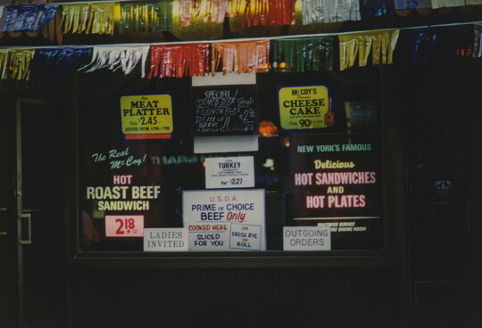 A color photograph of shop window, framed by colored fringed garlands, covered in advertisements for various foods for sale.