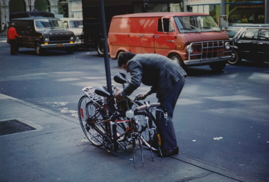 A color photograph of a man in a suit jacket bent over to lock or unlock a pair of bikes without front tires to a light post.