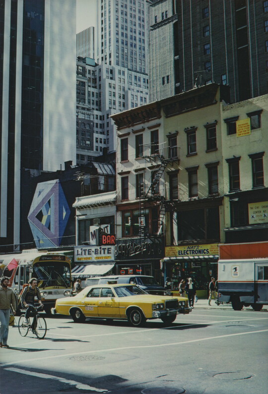 A color photograph of a busy city street with pedestrians, cyclists, cars, and busses.