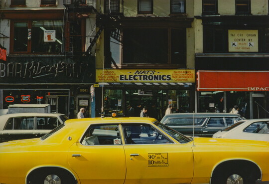 A color photograph of a yellow taxi in the foreground and other vehicles and storefronts in the background.