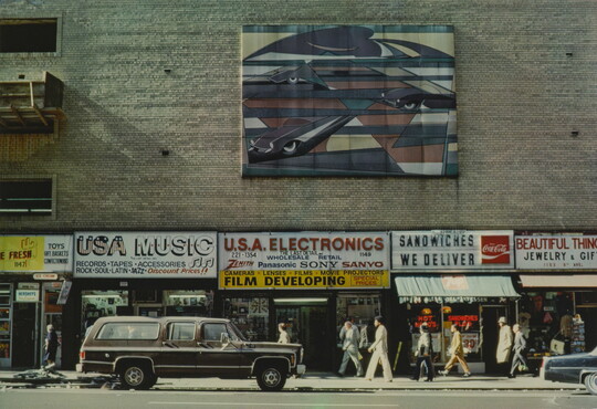 A color photograph of a brick building with colorful signs advertising products for different stores and a large work of art hanging above multiple stores.