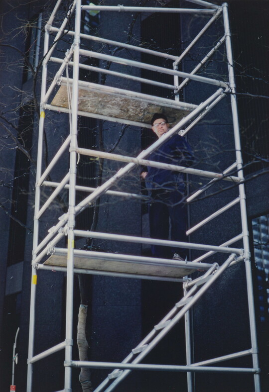 A color photograph of a White man in glasses standing on construction scaffolding looking down.