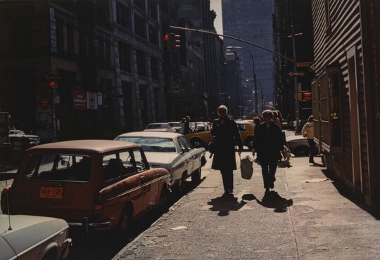 A color photograph of pedestrians on a city sidewalk next to parked cars, and cars passing through an intersection in the background.