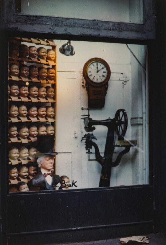 A color photograph of storefront window showing a shelf full of doll heads, a clock, and a piece of machinery.