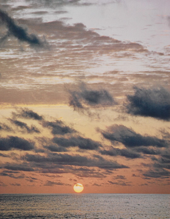 A color photograph of a sun just above an ocean horizon under wispy dark clouds.