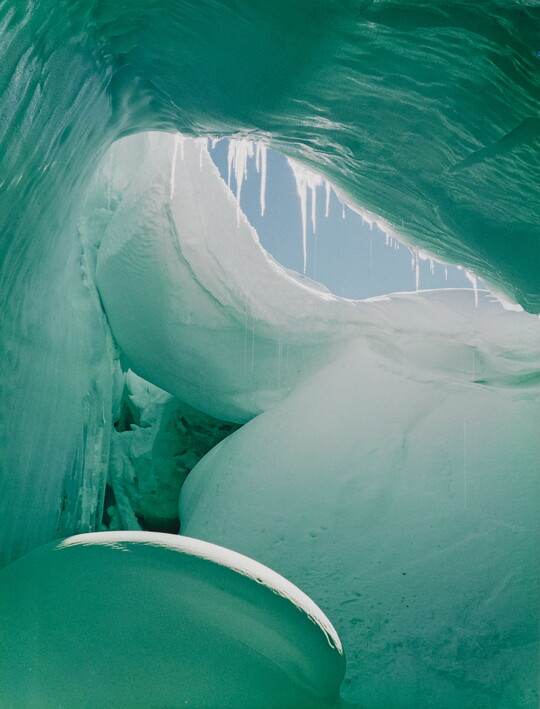 A color photograph from inside of a blue and green ice cave showing a sliver of blue sky.