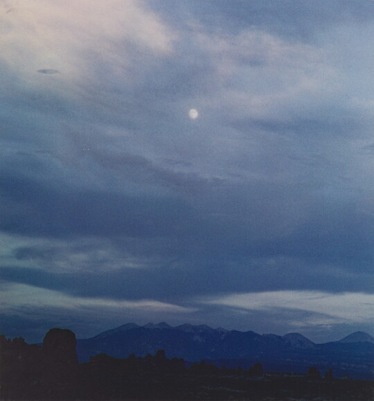 A color photograph of a mountain range below a full moon veiled behind thin dark clouds.