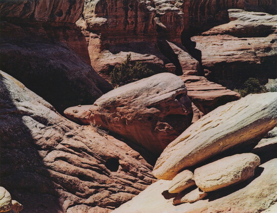 A color photograph of a red canyon with stark shadows and rocks balancing on a ledge in the foreground.