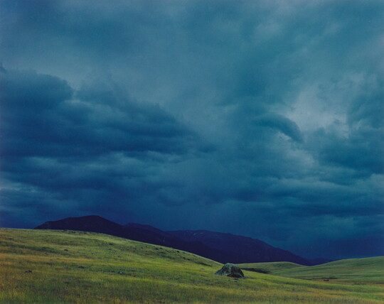 A color photograph of dark, heavy clouds above a mountain range beyond gentle green hills.