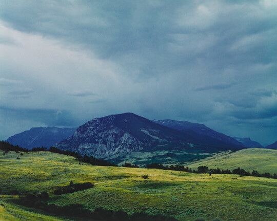 A color photograph of cloudy skies above a dark a mountain range beyond green fields in the foreground.