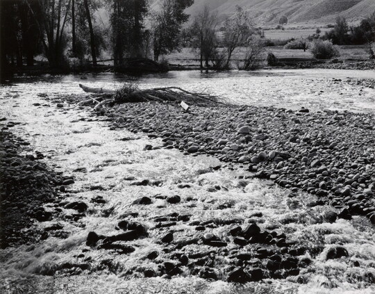 A black-and-white photograph of a mountain river bubbling through a gravel bar.