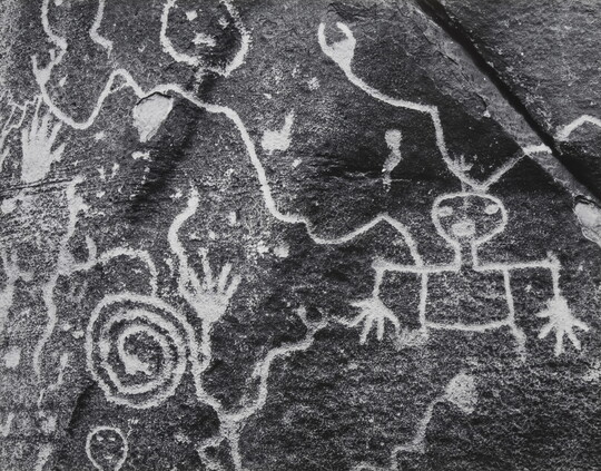 A close-up black-and-white photograph of symbols, several showing human forms, carved on a rock.