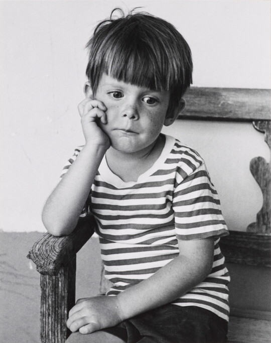 A black-and-white portrait photograph of a young White boy sitting in a wood chair resting his chin in his hand.