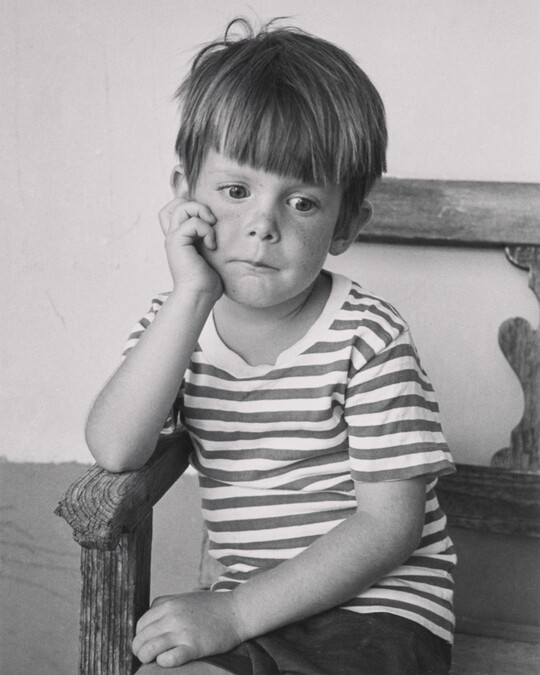 A black-and-white portrait photograph of a young White boy sitting in a wood chair resting his chin in his hand.