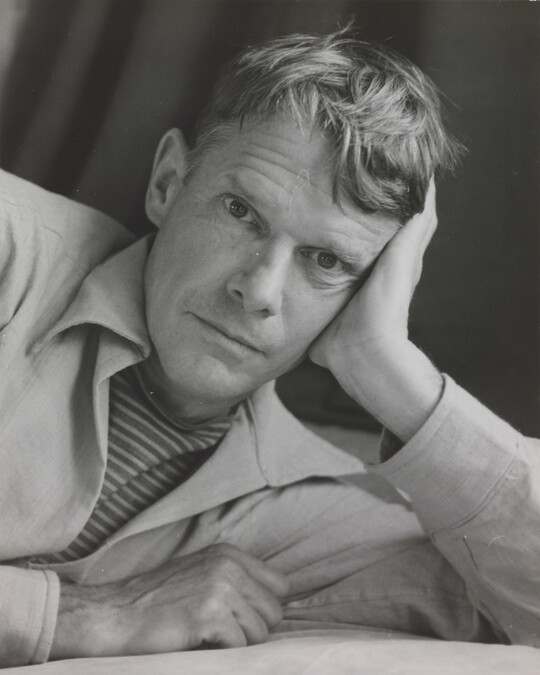 A black-and-white photograph portrait of a White man on his side, head resting on his hand, looking at the camera.