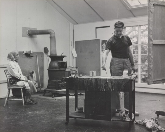 A black-and-white photograph of a White man working in an art studio and a White woman seated near him in a chair.