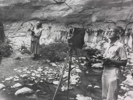 A black-and-white photograph of a White woman with a sketchbook and a White man with a camera next to a rock wall, both looking at something out of the frame.
