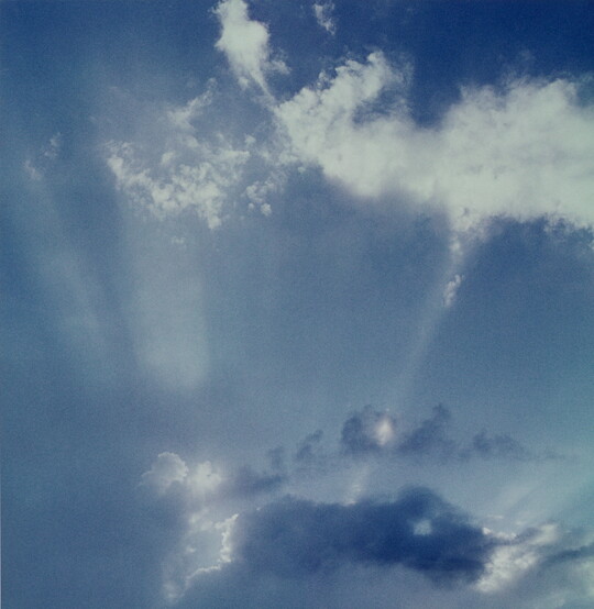 A color photograph of white clouds in a blue sky and sun rays that create white streaks.