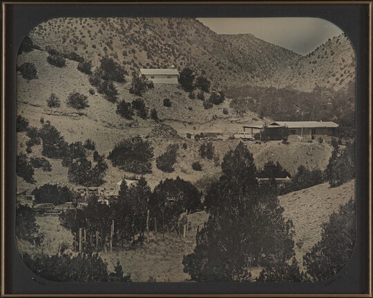 A black-and-white photograph of several buildings on a mountainside dotted with pine trees.